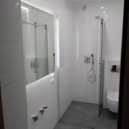 Nowoczesna, minimalistyczna łazienka z kabiną prysznicową typu walk-in, wykończona białymi płytkami z delikatnym wzorem, szara podłoga, widoczne zawory z oznaczeniami ciepłej i zimnej wody...