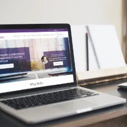 Laptop wyświetlający stronę internetową Klubu Inteligencji Finansowej, obok notatnik i myszka na drewnianym biurku.