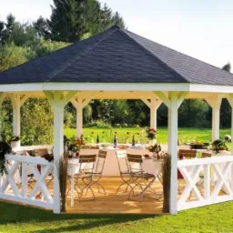 Biała altana ogrodowa z drewnianą podłogą i czarnym dachem, nakryty stół z zastawą i kwiatami wewnątrz, otoczona zielonym trawnikiem i drzewami.