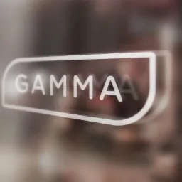 Podświetlane logo 'GAMMA' na szklanej powierzchni, rozmyte tło wnętrza, nowoczesny design z efektem głębi ostrości.