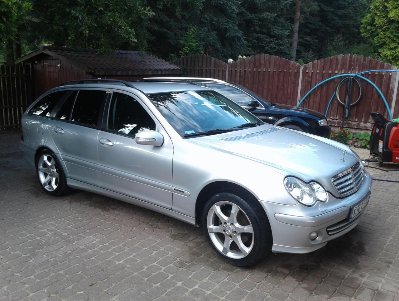 Srebrny samochód kombi marki Mercedes zaparkowany na kostce brukowej, widoczny wąż ogrodowy i myjka ciśnieniowa w tle.