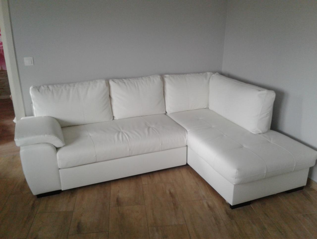 Biały, skórzany narożnik z poduszkami, ustawiony w rogu pokoju na drewnianej podłodze, widoczne delikatne przeszycia na siedzisku.