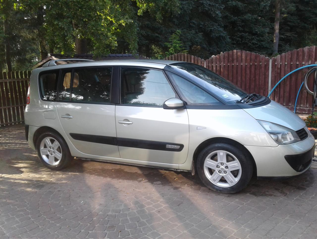 Srebrny minivan Renault Espace zaparkowany na brukowanym podjeździe, z widocznym ogrodzeniem z drewnianych sztachet i zielenią w tle.