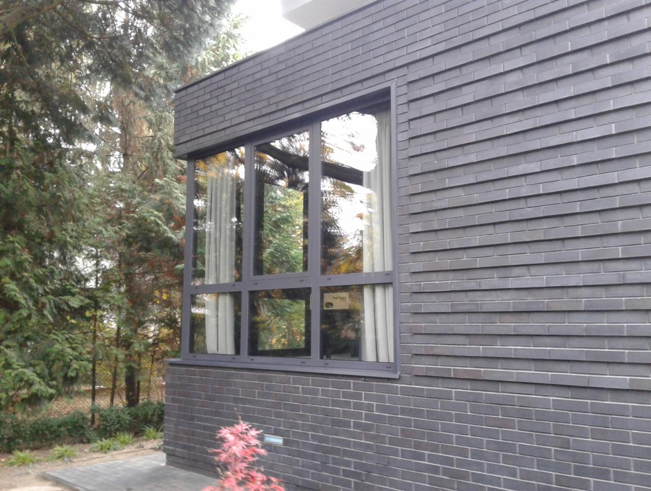 Szare cegły elewacyjne domu z dużym oknem z szarymi ramami, zasłoniętym firanką, na tle drzew i krzewów.