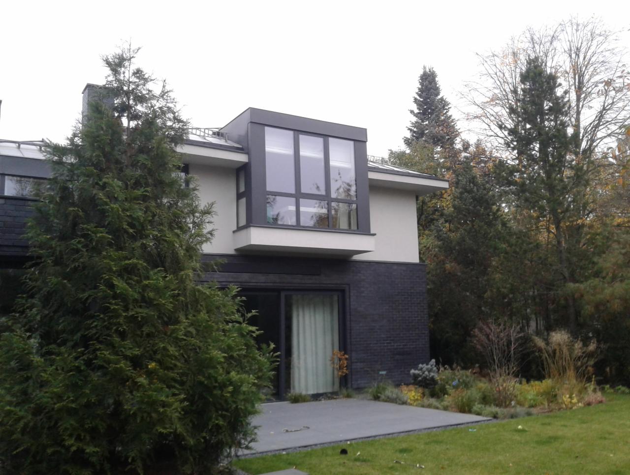 Nowoczesny dom z ciemnej cegły i dużymi oknami, otoczony zielenią, widoczny z perspektywy trawnika.