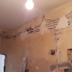 Wnętrze w trakcie remontu: ściany z bruzdami, w których ułożono kable elektryczne; widoczna puszka elektryczna i niezabezpieczone przewody; fragment rozdzielnicy elektrycznej na ścianie...