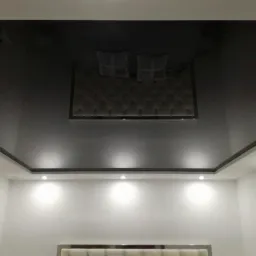 Błyszczący, czarny sufit z podwieszanym elementem i oświetleniem punktowym w nowoczesnej sypialni z białymi ścianami i widocznym fragmentem zagłówka łóżka.