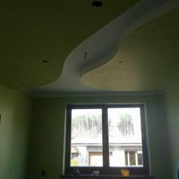 Usługi Remontowo-Budowlane Rafał Żukowski. Napinane sufity - Pokój z zielonymi ścianami i falistym, białym elementem dekoracyjnym na suficie, widoczne otwory na oświetlenie, okno z widokiem na zewnątrz i grzejnik pod nim.
