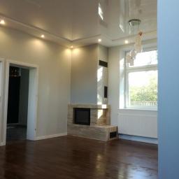 Usługi Remontowo-Budowlane Rafał Żukowski. Napinane sufity - Jasne, przestronne wnętrze z błyszczącą podłogą, nowoczesnym kominkiem z kamienia, gładkim sufitem podwieszanym z oświetleniem punktowym i wiszącą lampą, widok na zieleń za oknem.