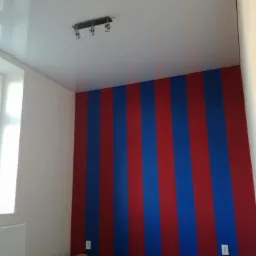 Pokój po malowaniu ścian w pionowe pasy czerwono-niebieskie; widoczny fragment sufitu z lampą i biała ściana z oknem.