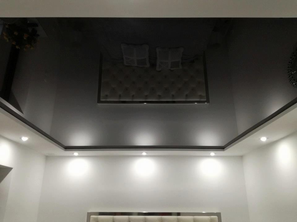 Błyszczący, czarny sufit z podwieszanym elementem i oświetleniem punktowym w nowoczesnej sypialni z białymi ścianami i widocznym fragmentem zagłówka łóżka.