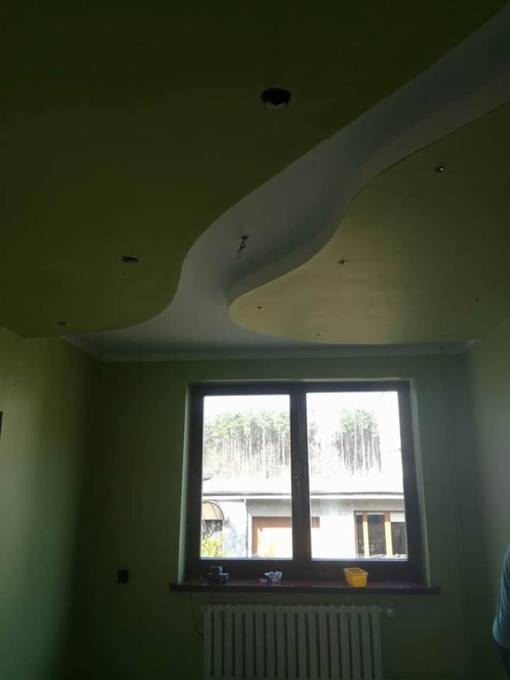 Pokój z zielonymi ścianami i falistym, białym elementem dekoracyjnym na suficie, widoczne otwory na oświetlenie, okno z widokiem na zewnątrz i grzejnik pod nim.