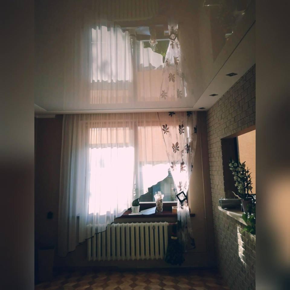 Jasne wnętrze pokoju w Ostrowi Mazowieckiej z dużym oknem, białymi zasłonami z motywem liści, błyszczącym sufitem i ceglaną ścianą po prawej stronie.