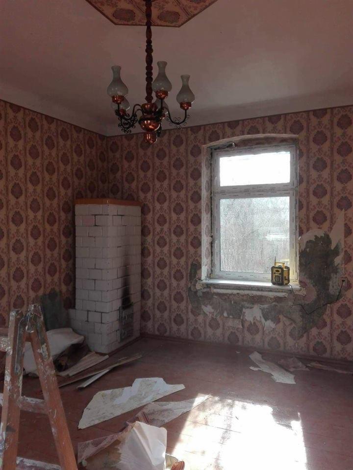 Pokój w trakcie demontażu tapety, widoczny fragment ściany z usuniętą tapetą, drabina malarska, resztki tapety na podłodze, piec z białej cegły i okno z radiem na parapecie.