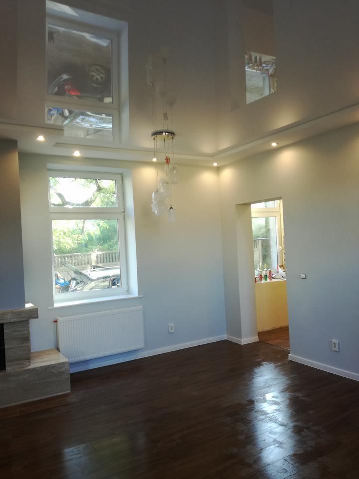 Jasne, przestronne wnętrze po remoncie z ciemną, błyszczącą podłogą, białymi ścianami i nowoczesnym oświetleniem punktowym oraz lampą wiszącą. W tle widoczne okno z widokiem na zieleń.