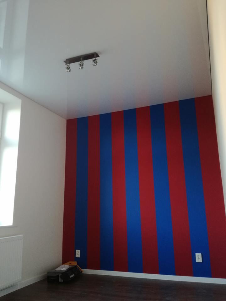 Pokój po malowaniu ścian w pionowe pasy czerwono-niebieskie; widoczny fragment sufitu z lampą i biała ściana z oknem.