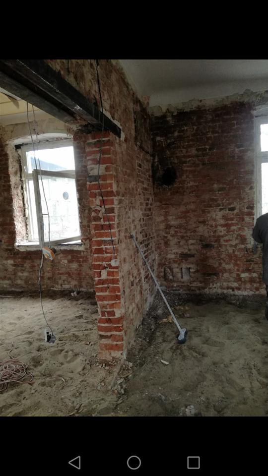 Wnętrze w trakcie remontu: odsłonięta cegła, metalowa belka stropowa, piasek na podłodze, widoczne przewody elektryczne, okno z widokiem na zewnątrz.