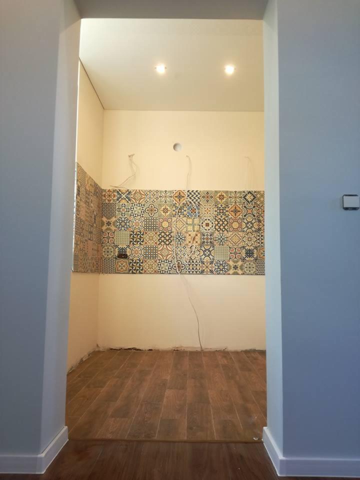 Wnętrze łazienki w trakcie remontu, z położonymi płytkami dekoracyjnymi na ścianie, widocznymi przewodami elektrycznymi i drewnopodobną podłogą, uchwycone z perspektywy wejścia.