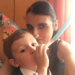 Kobieta trzyma na rękach małego chłopca w stroju galowym i czapeczce urodzinowej, dziecko dmucha w gwizdek urodzinowy, pomarańczowy balon w tle.