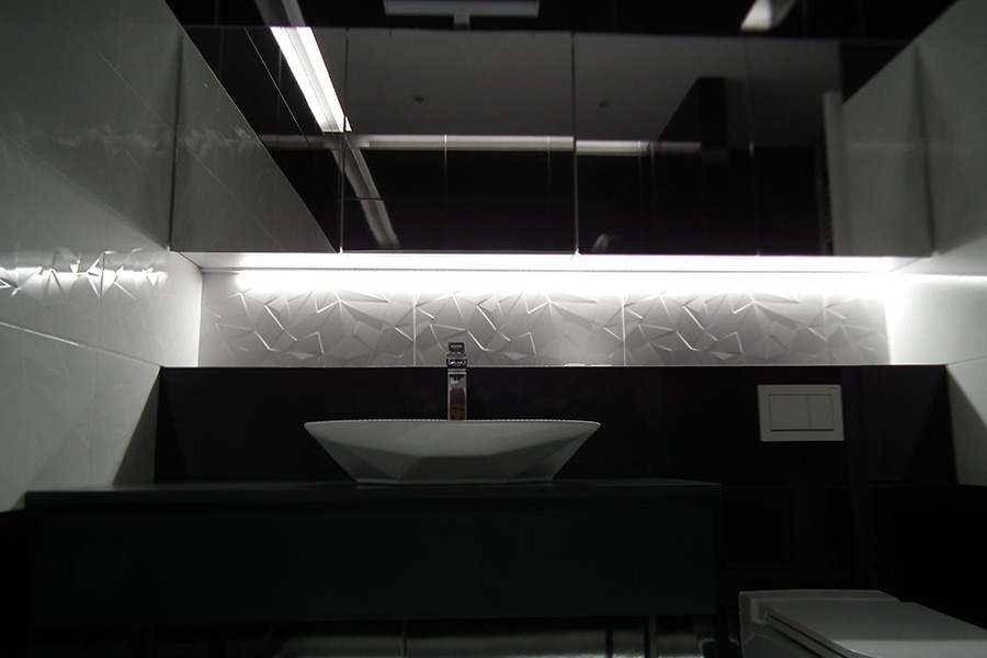 Nowoczesna łazienka w Katowicach z umywalką w kształcie diamentu, chromowaną baterią i podświetlanymi trójwymiarowymi białymi płytkami na ścianie, odbijającymi się w czarnych, lakierowanych szafkach.