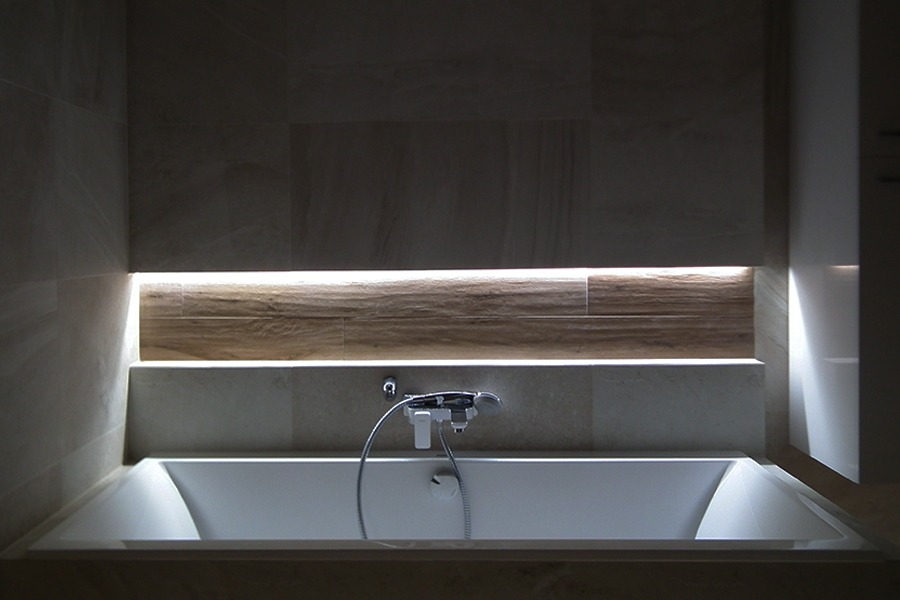 Wanna obudowana płytkami z podświetleniem LED w niszy, bateria wannowa zamontowana na ścianie, minimalistyczny design łazienki.