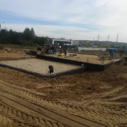 Wykopany i przygotowany teren pod budowę domu, zalane fundamenty z wystającymi prętami zbrojeniowymi, w tle pracujący robotnicy i sprzęt budowlany.