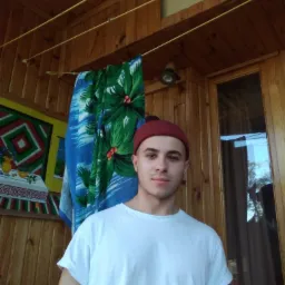 Młody mężczyzna w białej koszulce i bordowej czapce pozuje na tle drewnianej werandy z kolorową dekoracją i ręcznikiem.