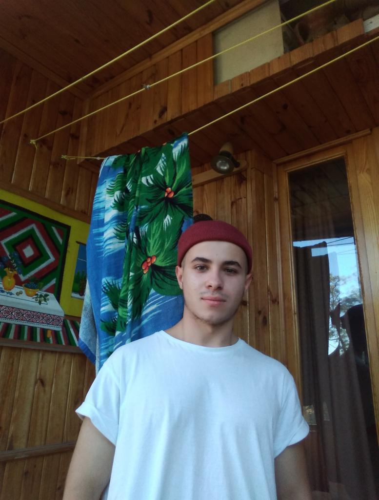 Młody mężczyzna w białej koszulce i bordowej czapce pozuje na tle drewnianej werandy z kolorową dekoracją i ręcznikiem.