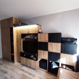 Pańczuk Meble Łukasz Pańczuk - Zabudowa meblowa w salonie z szafą, szafką RTV i regałem z czarno-drewnianymi modułami. Na górze regału drukarka. Na szafie zabawka. Drewniana podłoga.