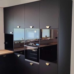 Pańczuk Meble Łukasz Pańczuk - Czarna zabudowa kuchenna z piekarnikiem i mikrofalówką, złote uchwyty, drewniany blat i podłoga. Widoczne odbicia w szklanych frontach szafek.