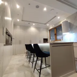 Nowoczesna kuchnia na wymiar w Czarna Białostocka: białe fronty szafek, wyspa z podświetleniem LED i czarne hokery. Marmurowe wykończenie ścian i podłogi.