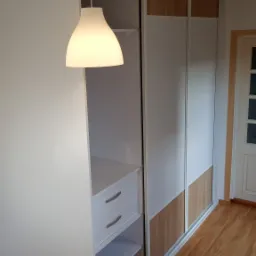 Biała szafa wnękowa z przesuwnymi drzwiami, z elementami w kolorze drewna, oświetlona wiszącą lampą, widok na półki i szuflady.