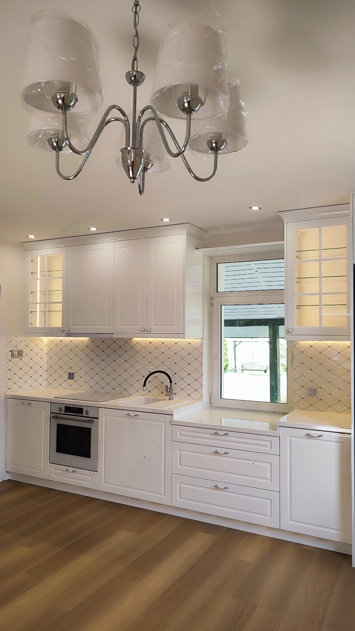 Biała kuchnia z frezowanymi frontami, oświetleniem LED podszafkowym i dekoracyjną mozaiką nad blatem. Lampa z abażurami w folii ochronnej. Widok na zewnątrz przez okno.