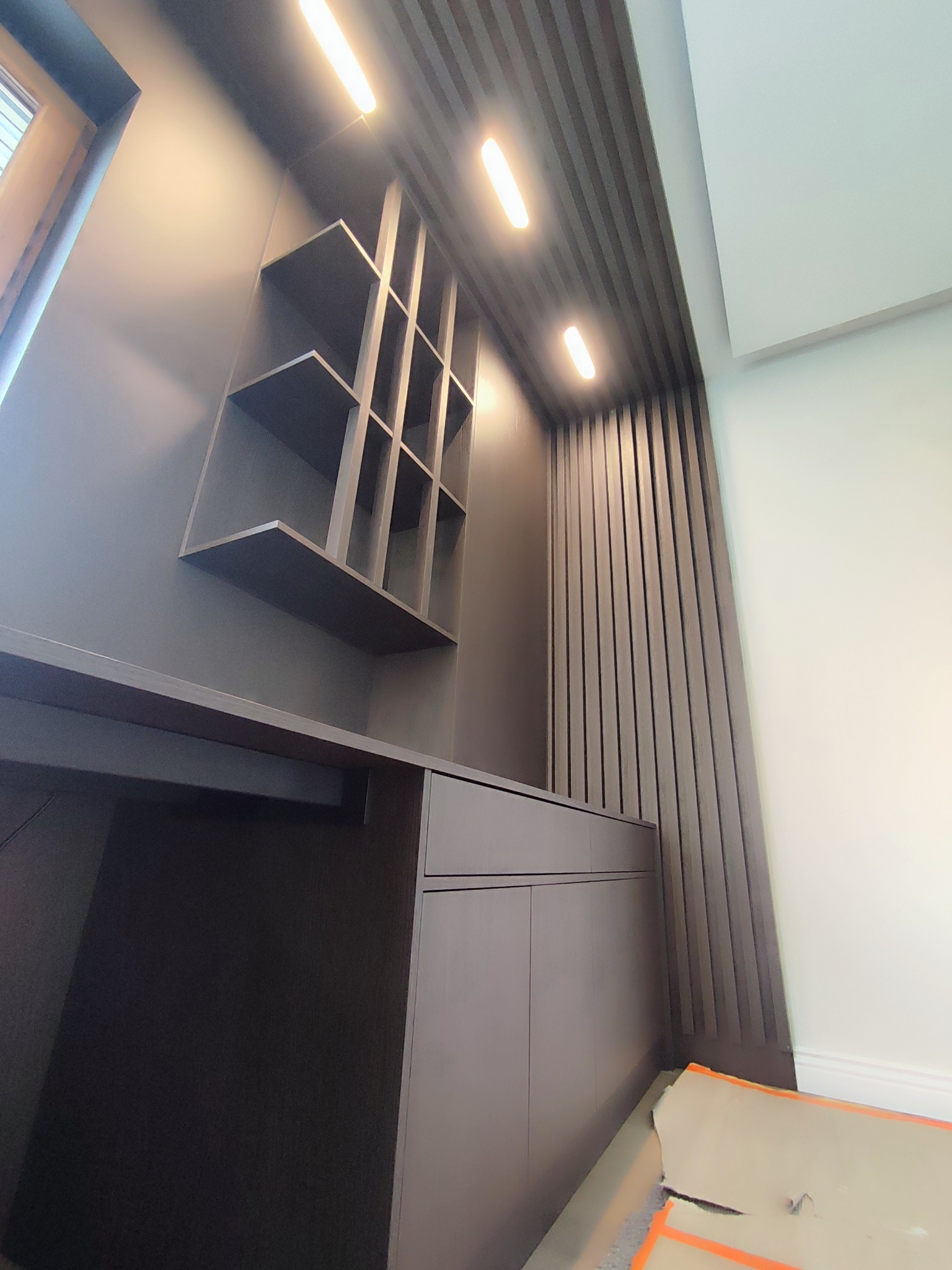 Nowoczesne biurko z szafkami i półkami w zabudowie, oświetlenie punktowe, dekoracyjne panele ścienne w pionowe pasy. Ujęcie z dołu podkreśla wysokość zabudowy.