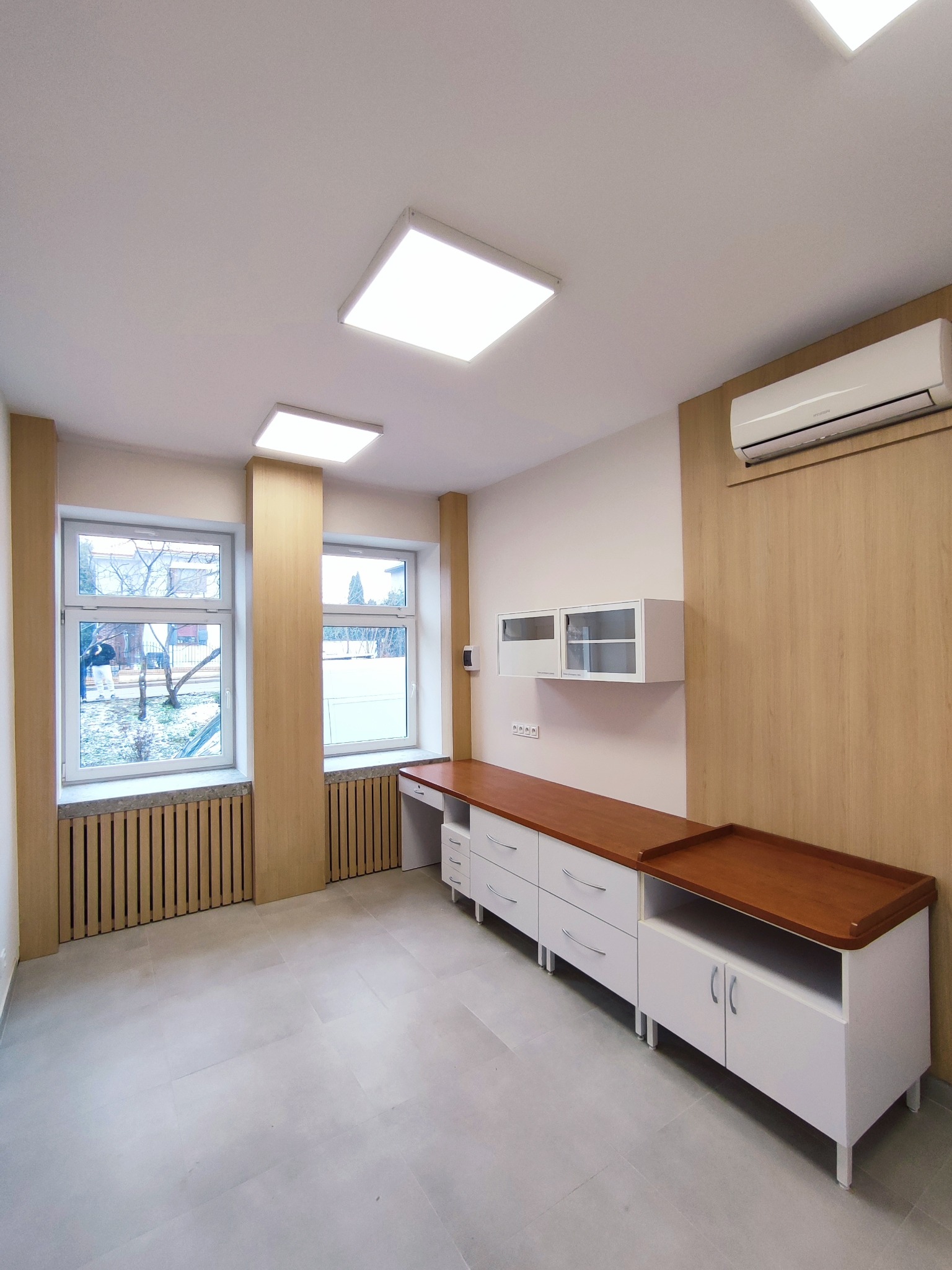 Nowoczesne biuro z zabudową meblową: białe szafki z drewnianym blatem, wiszące szafki, klimatyzacja. Jasne wnętrze z widokiem na zewnątrz.