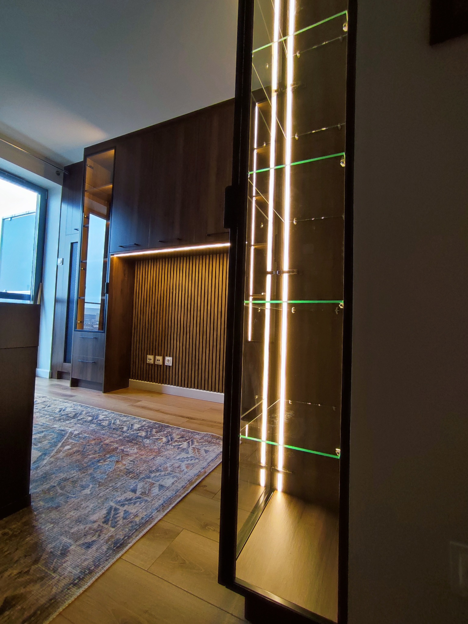 Nowoczesna, ciemna zabudowa meblowa z podświetlaną szafą szklaną i drewnianą ścianą z pionowymi panelami. Dywan w stylu vintage na drewnianej podłodze.