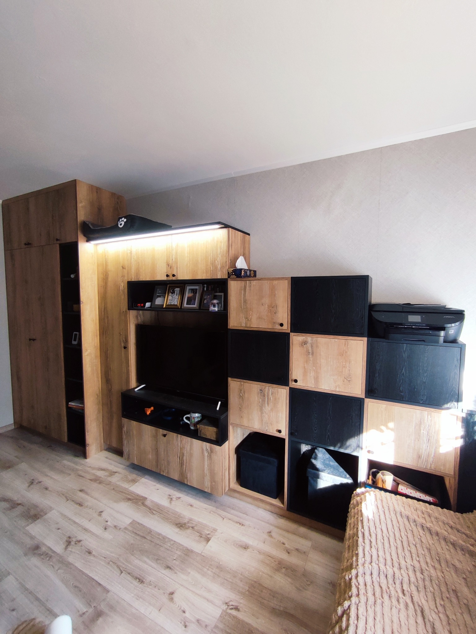 Zabudowa meblowa w salonie z szafą, szafką RTV i regałem z czarno-drewnianymi modułami. Na górze regału drukarka. Na szafie zabawka. Drewniana podłoga.