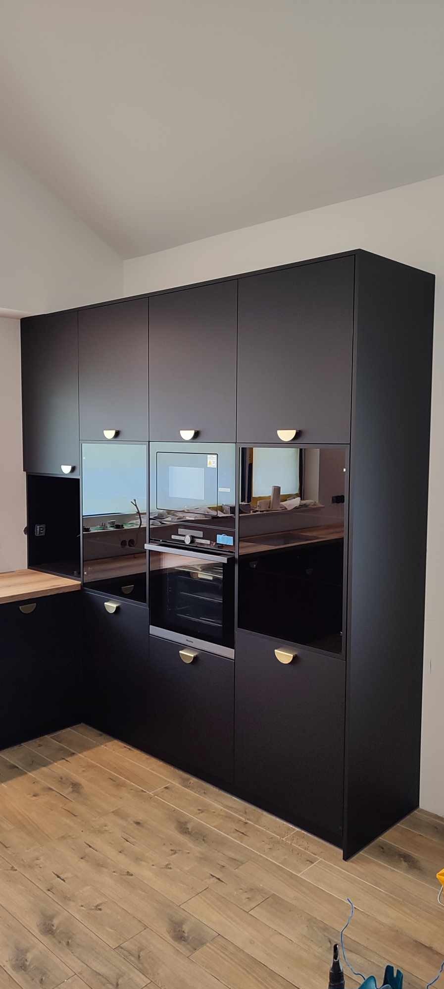 Czarna zabudowa kuchenna z piekarnikiem i mikrofalówką, złote uchwyty, drewniany blat i podłoga. Widoczne odbicia w szklanych frontach szafek.