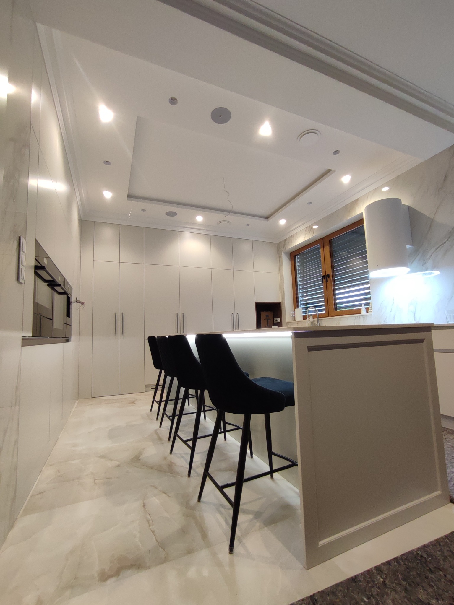 Nowoczesna kuchnia na wymiar w Czarna Białostocka: białe fronty szafek, wyspa z podświetleniem LED i czarne hokery. Marmurowe wykończenie ścian i podłogi.