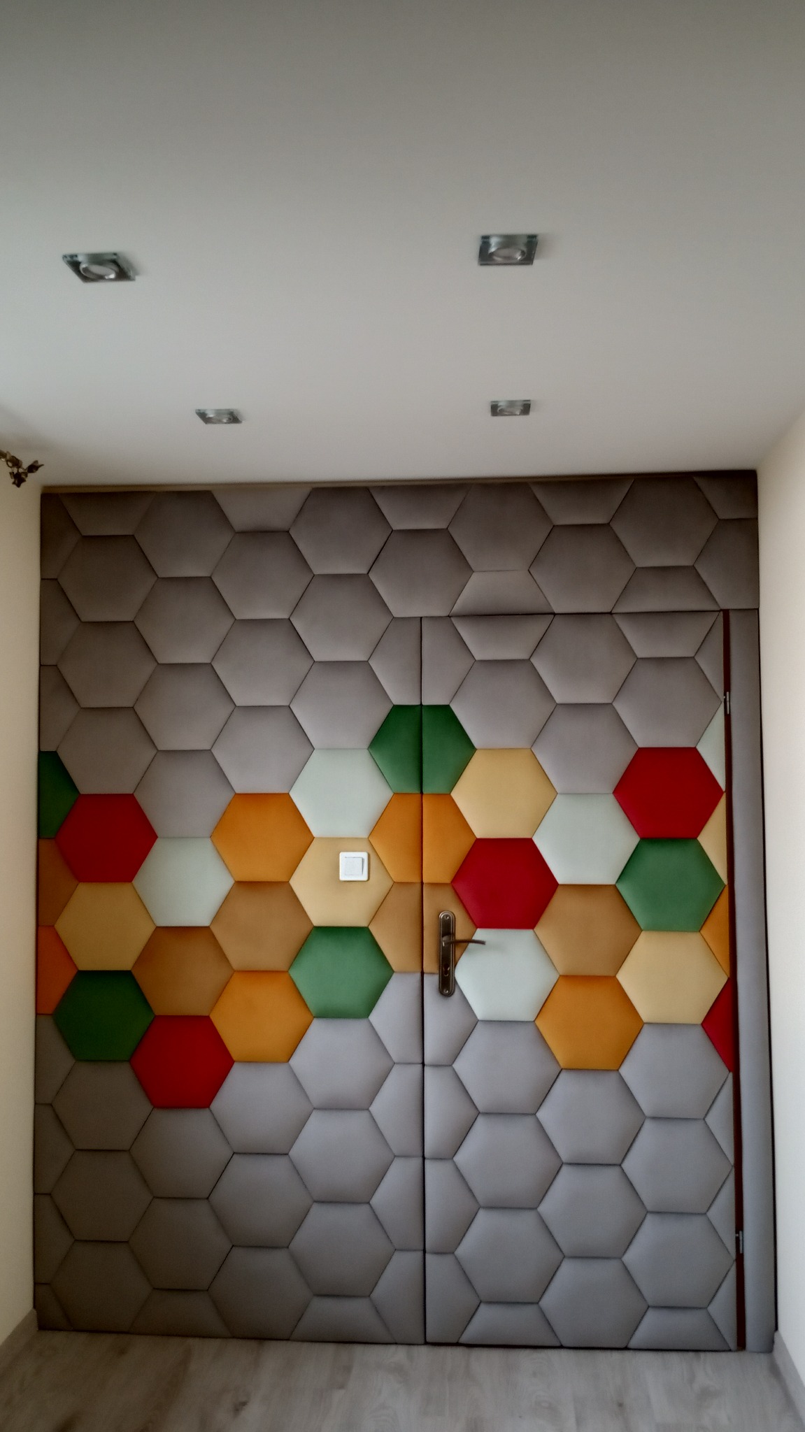 Dwuskrzydłowe drzwi wewnętrzne obite panelami tapicerowanymi w kształcie szarych heksagonów, z kolorowymi akcentami w odcieniach czerwieni, zieleni i żółci, tworzącymi wzór na dolnej części drzwi.