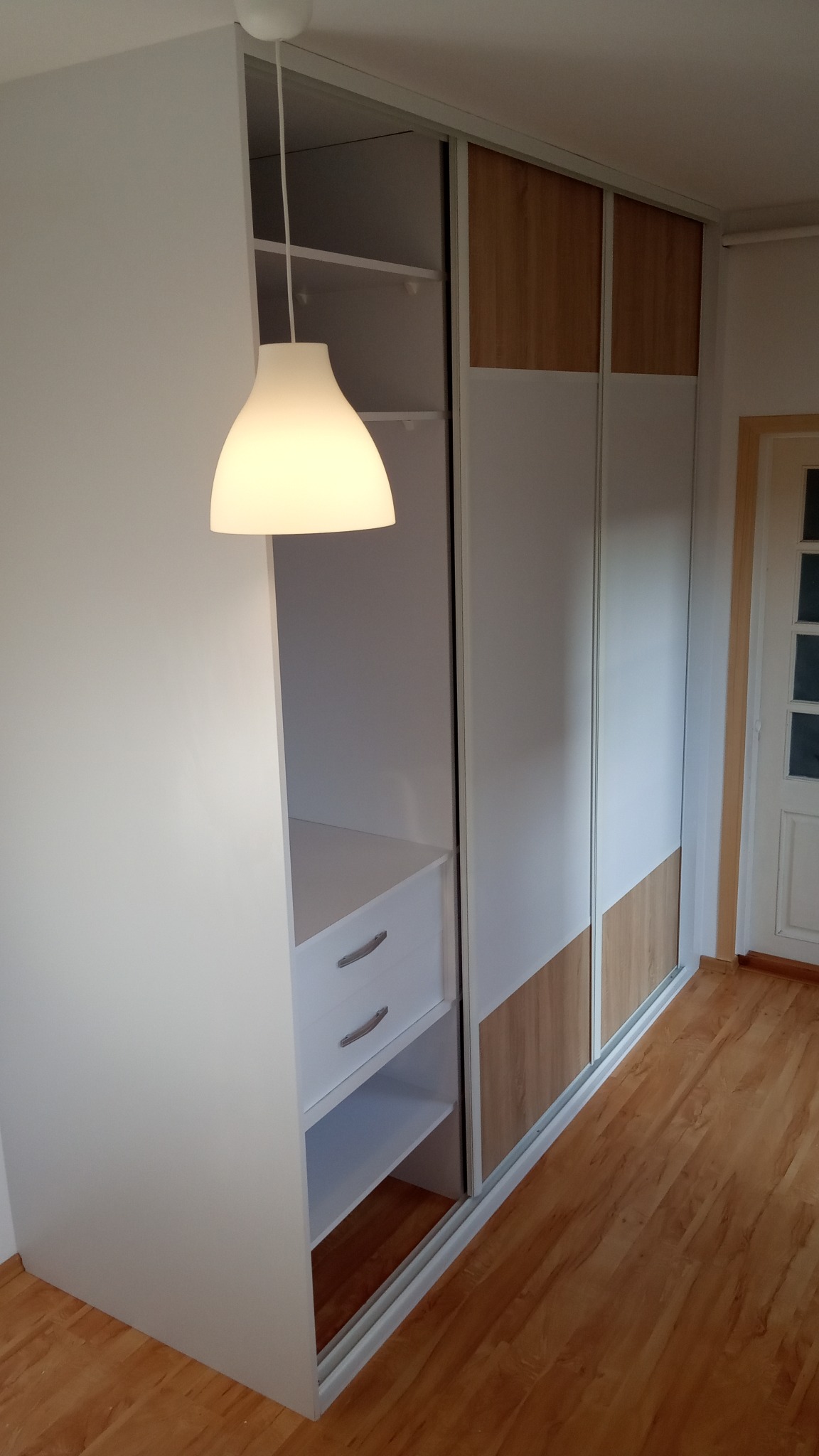 Biała szafa wnękowa z przesuwnymi drzwiami, z elementami w kolorze drewna, oświetlona wiszącą lampą, widok na półki i szuflady.