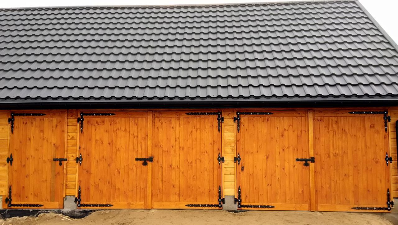 Drewniana konstrukcja z czterema bramami garażowymi, każda z czarnymi, ozdobnymi zawiasami i zamkami, pod czarnym dachem z blachy trapezowej.