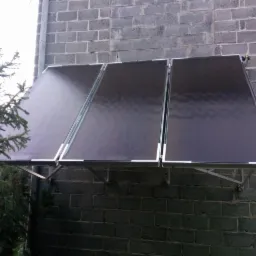 Trzy kolektory słoneczne zamontowane na szarej, ceglanej ścianie budynku, widoczne elementy instalacji i fragment zieleni.