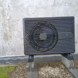 Czarna pompa ciepła marki FOX Air, zamontowana na betonowych wspornikach, z widoczną elewacją budynku w tle i kamieniami dekoracyjnymi u podstawy.