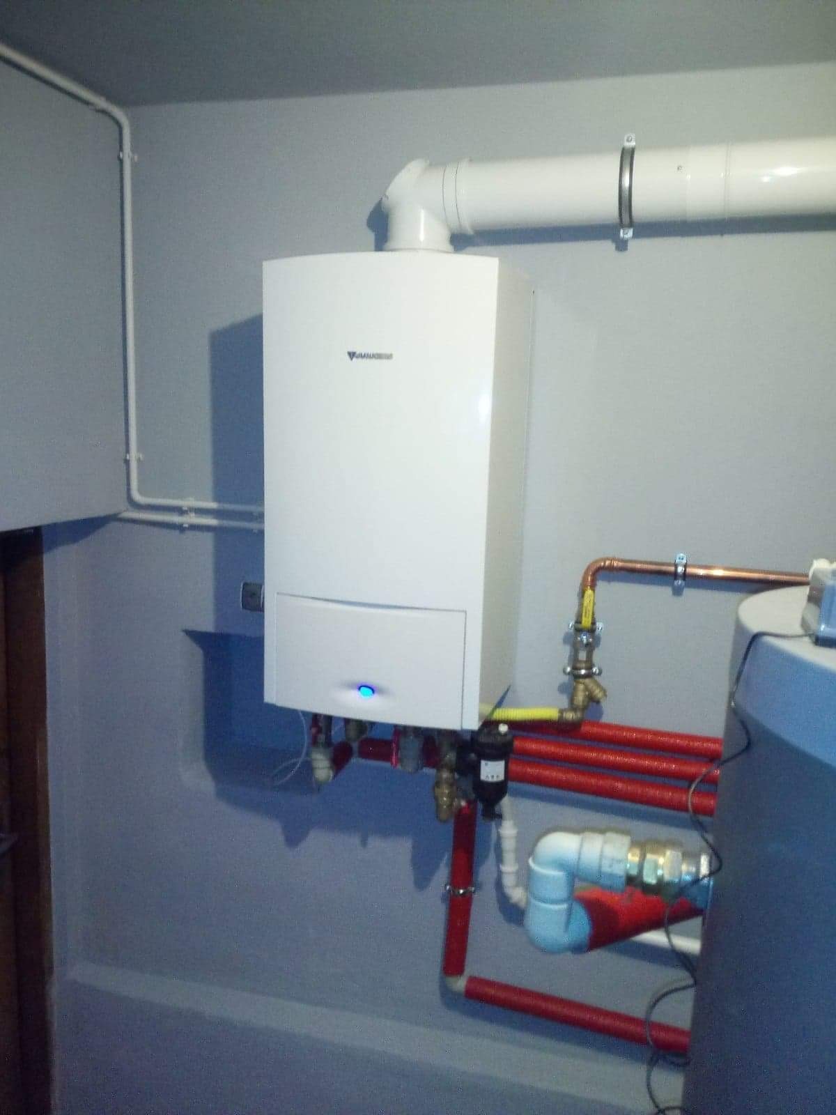 Biały piec gazowy Junkers zamontowany na szarej ścianie, z widocznymi czerwonymi rurami instalacji grzewczej i białym przewodem odprowadzającym spaliny