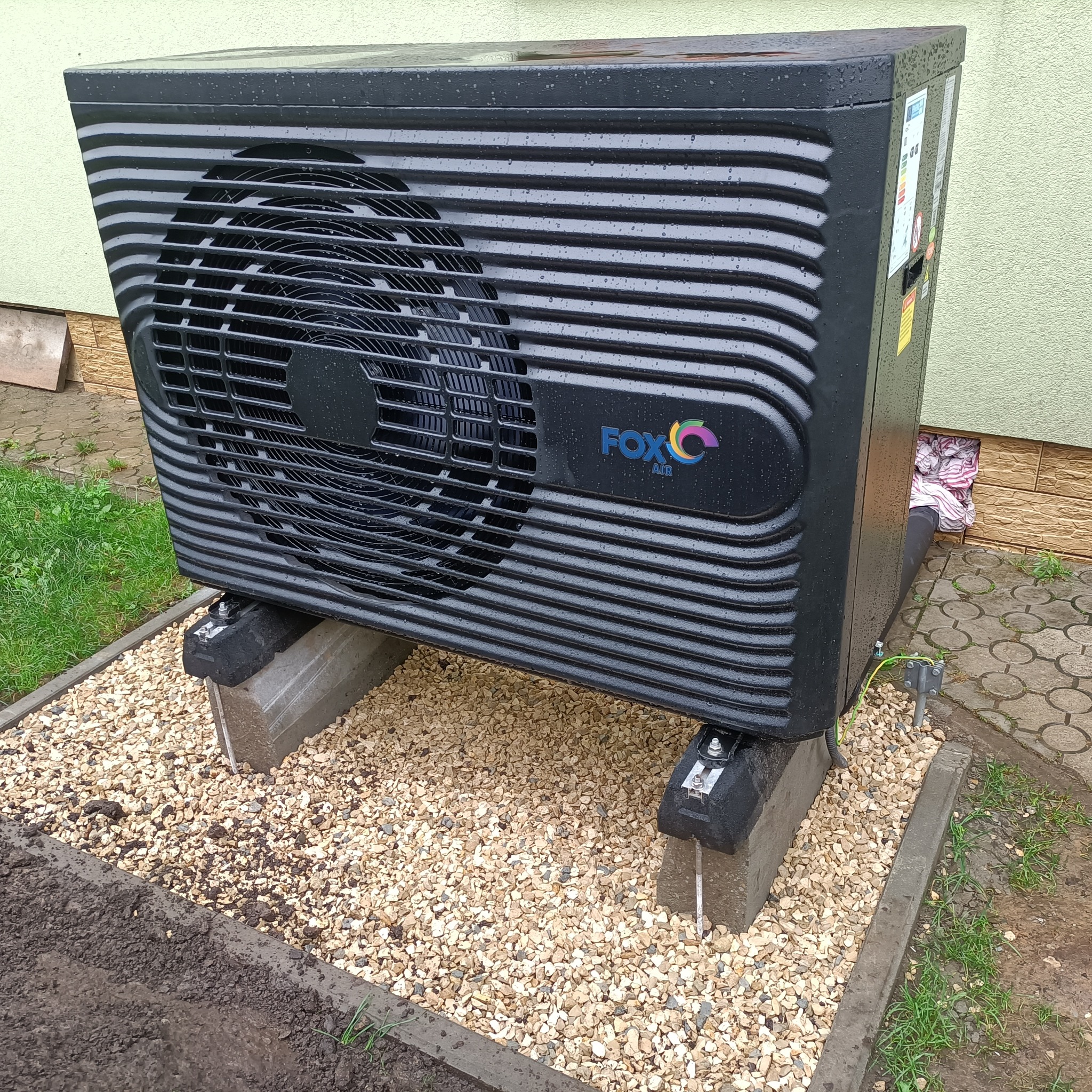 Czarna pompa ciepła marki Fox Air, ustawiona na betonowych blokach na żwirowym podłożu, widoczne krople deszczu na obudowie, z tyłu fragment ściany budynku.
