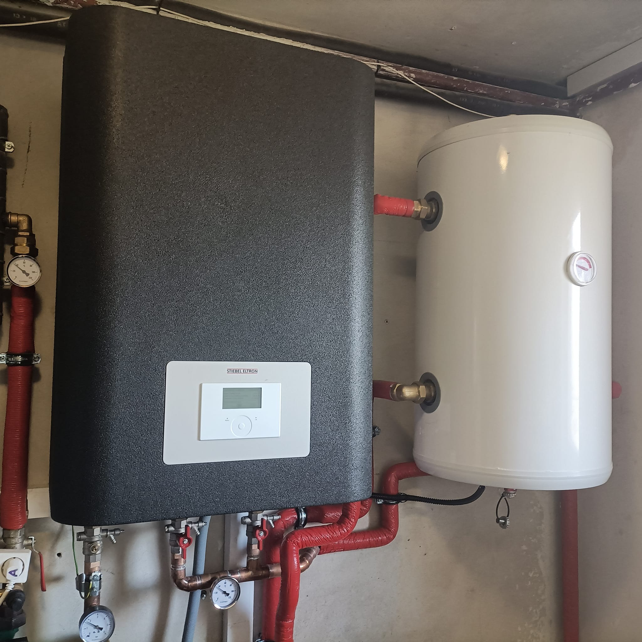 Czarny panel sterowania pompy ciepła Stiebel Eltron, obok biały zasobnik ciepłej wody, rury miedziane z czerwoną izolacją, manometry i zawory, instalacja przy szarej ścianie.