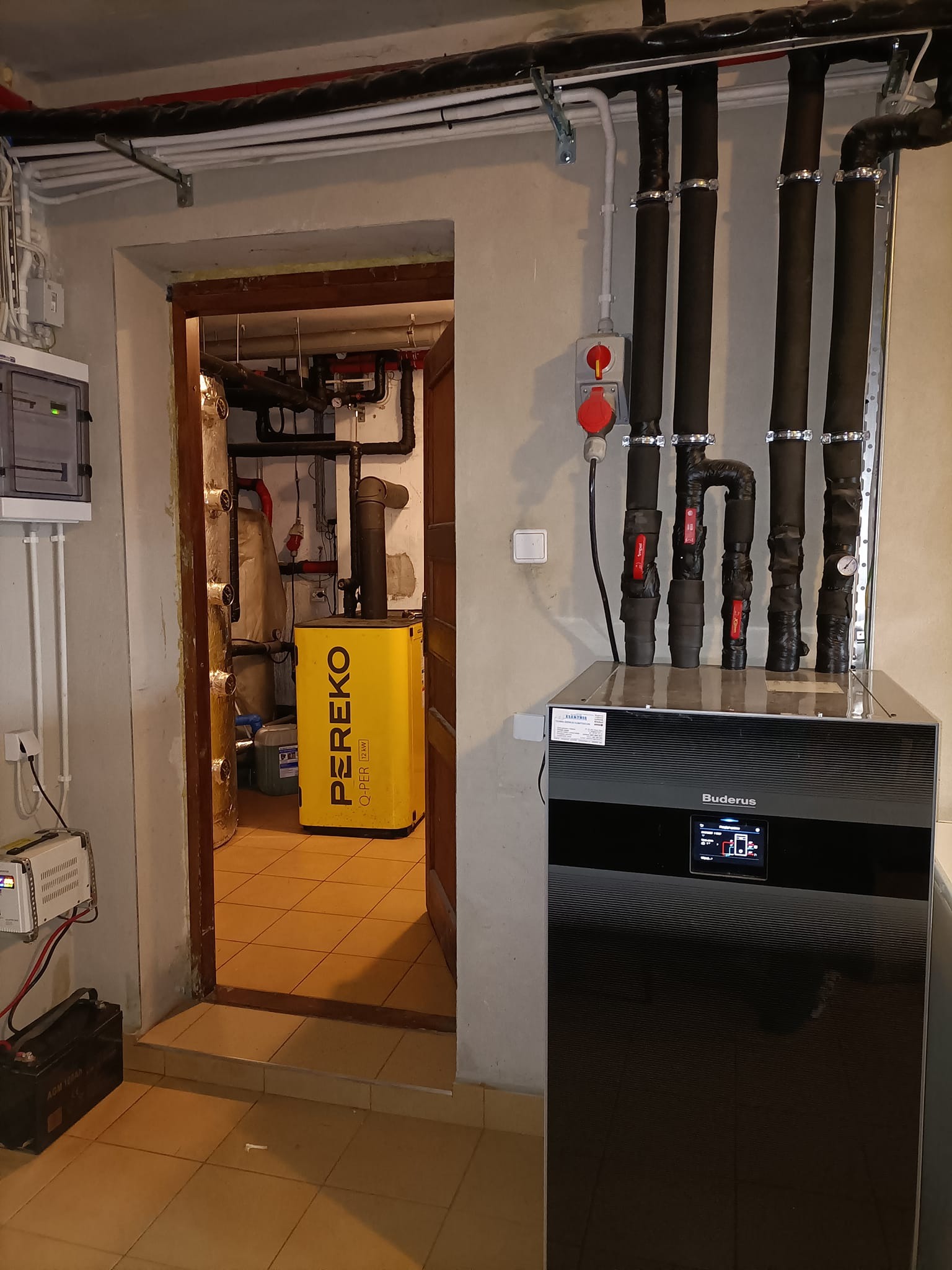 Instalacja grzewcza w piwnicy: czarny piec Buderus z wyświetlaczem, widoczne rury izolowane termicznie, w tle przez otwarte drzwi żółty piec Pereko.
