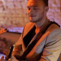 Młody mężczyzna w szarym garniturze gra na gitarze elektrycznej, oświetlony ciepłym światłem na tle ceglanej ściany.