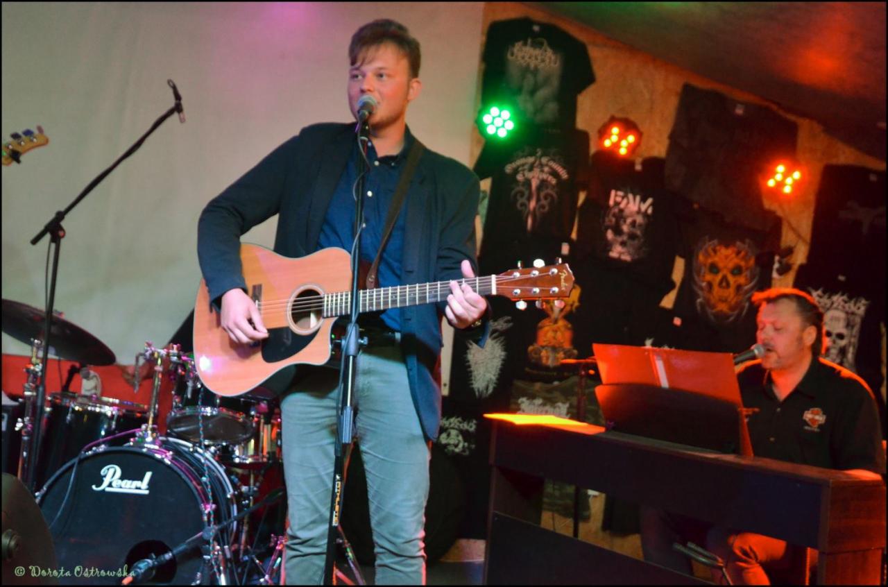 Wokalista z gitarą akustyczną na scenie podczas występu, w tle perkusja, klawiszowiec i ściana z koszulkami z motywami metalowymi.